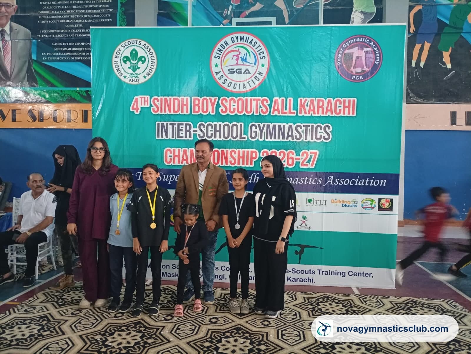 Nova gymnasts wearing their medals with coaches in front of the 4th Sindh Boy Scouts All Karachi Inter-School Gymnastics Championship 2026–27 banner
